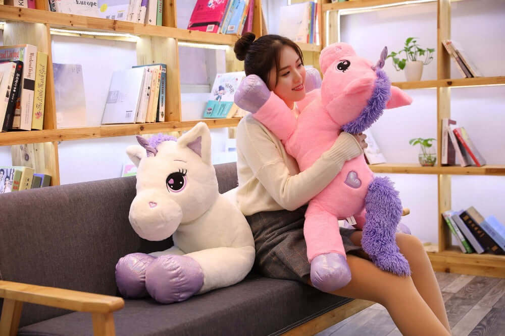 Smiling woman cuddling a soft unicorn pillow toy while sitting on a couch surrounded by books, perfect for toddlers and children.